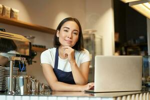 Portrait of smiling korean woman, barista in coffee shop, standing at counter with laptop, smiling and looking confident, self-employed female entrepreneur in her own coffee shop photo