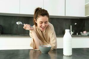 Portrait of young beautiful woman in bathrobe, eating cereals for breakfast, leans on kitchen worktop, looking at her morning meal photo