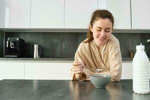 Portrait of happy young woman leans on kitchen worktop and eating cereals, has milk and bowl in front of her, having her breakfast, wearing bathrobe photo