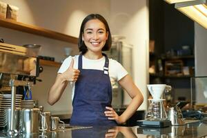 sonriente asiático barista, niña en café uniforme, muestra bueno firmar, recomienda su café, aprobar nuevo sabores, en pie en delantal foto