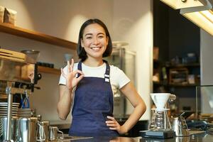 retrato de sonriente coreano barista, niña a el encimera, usa azul delantal, trabajos en café comercio, muestra bueno firmar foto