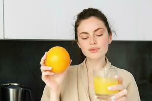 Portrait of beautiful, healthy young woman with orange in hand, drinks freshly squeezed juice from glass, posing in kitchen, enjoys her morning at home photo