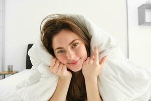 Portrait of gorgeous smiling woman, lying in bed covered in duvet, has messy hair, looking happy, relaxing in her bedroom, spending time in hotel room in morning photo