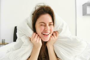 Portrait of gorgeous smiling woman, lying in bed covered in duvet, has messy hair, looking happy, relaxing in her bedroom, spending time in hotel room in morning photo