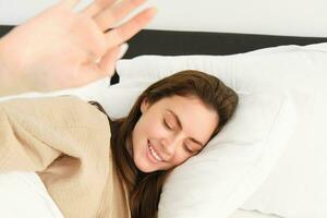 Portrait of gorgeous girlfriend stretching hand, blocking camera, lying in bed and resting on pillow, smiling while waking up from someone photo