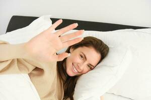 Portrait of beautiful brunette woman in pyjamas, stretches hand, waving at camera and smiling. Girlfriend being waken up by partner, lying in bed, enjoying her morning photo