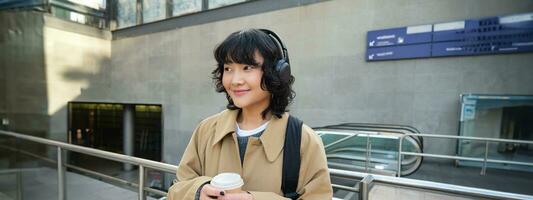 Portrait of young student, girl in headphones, drinks coffee, stands on street with backpack, commutes to university or college, smiles happily photo