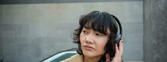 Portrait of complicated young korean girl in headphones, frowns and looks disappointed while stands and waits for public transport on street photo