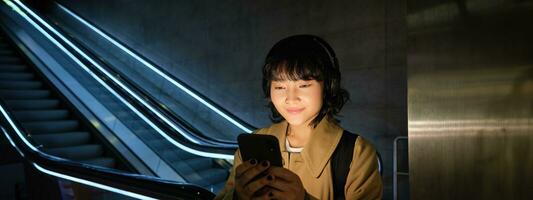 Portrait of young smiling asian girl, stands near escalator and waits for someone, reads message, checks map on smartphone, listens music in headphones photo