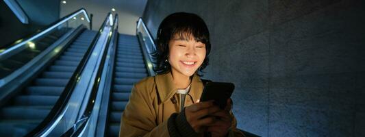 Smiling korean girl going down escalator in dark, holding mobile phone, using smartphone app, listening music, commuting in city photo