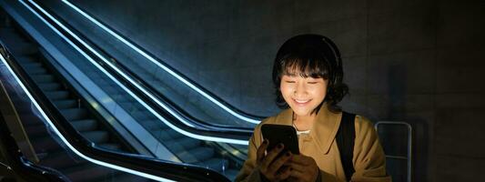 Happy asian woman in subway, standing near escalator, holding smartphone and listening music in headphones, smiling pleased photo