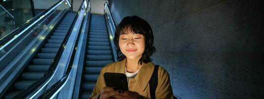 Portrait of happy beautiful asian girl, standing with smartphone on escalator, going down to metro station, commuting via tube photo