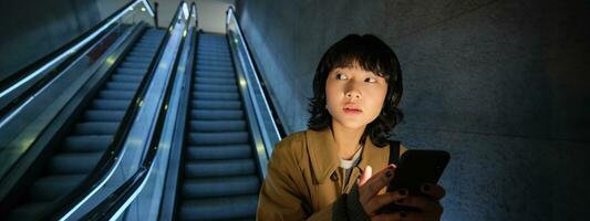 urbano estilo de vida y gente. retrato de coreano niña en auriculares, sostiene teléfono inteligente, mira preocupado y tenso mientras yendo abajo escalera mecánica en oscuro foto
