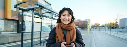 retrato de coreano mujer en invierno chaqueta, en pie con teléfono inteligente, esperando para autobús en detener, mirando a móvil teléfono aplicación comprobación público transporte solicitud foto