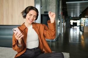 Close up portrait of happy employee, young office manager sitting on floor in break room, resting in co-working space, wearing wireless headphones, holding smartphone and dancing photo