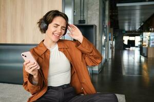 Portrait of modern young woman, office worker in lounge zone, dancing and sitting on floor of break room, enjoying her lunch, singing and listening music on smartphone photo