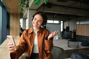 Image of smiling young woman, company employee, sitting in office with smartphone, demonstrating creative co-working space to people online, showing smth, video chats on mobile phone photo