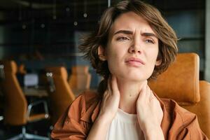 Image of woman with tired face, sits with laptop in office, feels tension in neck, pain in muscles after working all day at workplace photo