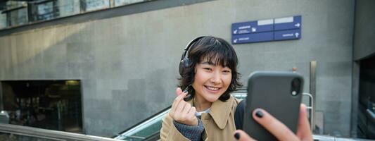 Cute and stylish Korean girl, wears headphones, takes selfie on smartphone, tourist records video or makes a photo, stands on street photo