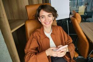 Image of young professional entrepreneur, woman in an office, sitting in chair in an office, using mobile phone, messaging a co-worker photo