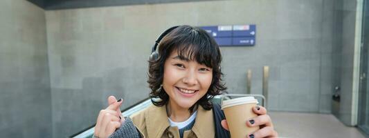 Portrait of stylish asian woman in headphones, drinks coffee to go and smiles, enjoys cappuccino while commutes, stands on street photo