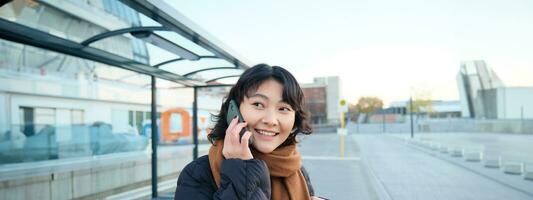 Beautiful smiling korean girl, waiting on bus stop, using public transport, talking on mobile phone, going somewhere in city photo
