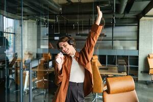 Carefree woman in office dancing and having fun, listening music in headphones, holding mobile phone, enjoys lunch break photo