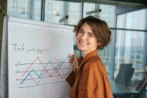 Portrait of young businesswoman, digital nomad standing with marker near board in office, drawing a diagram, giving presentation in front of team, smiling at camera photo