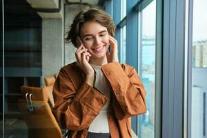 Portrait of working woman, social media manager in office, talking on mobile phone, having conversation on telephone, standing next to the window photo