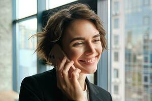 Close up shot of businesswoman talking over mobile phone and smiling, standing near office window and looking happy photo