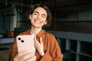 Portrait of woman in office, wearing casual clothes, standing with smartphone and laughing over funny video, talks to someone on video chat app photo