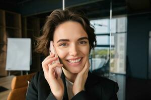 Close up portrait of smiling saleswoman, office manager stands in conference room, answers phone call, talking on mobile telephone with someone, receives great news and rejoices photo