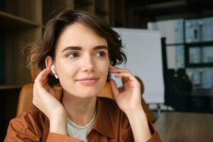 Close up portrait of woman in wireless earphones, smiling and sitting in office, listening to music photo