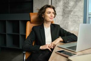 Portrait of successful woman in office chair, working for company, sitting near laptop and looking outside window with satisfied smiling face photo