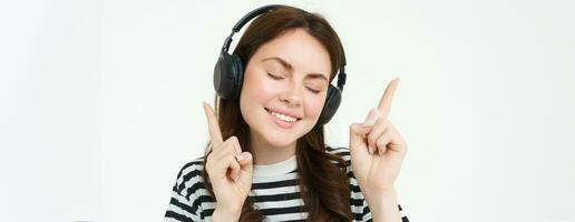 retrato de contento estudiante chica, escuchando música en auriculares, bailando y señalando dedos a Copiar espacio, promoción texto, aislado en contra blanco antecedentes foto