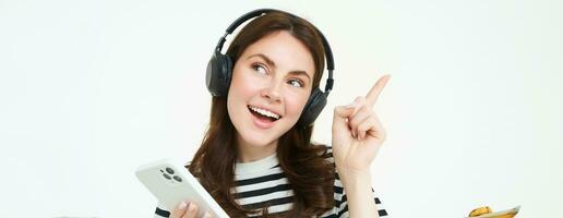 contento hermosa mujer, escuchando música en inalámbrico auriculares, participación móvil teléfono, señalando izquierda a Copiar espacio, demostración anuncio publicitario, blanco antecedentes foto