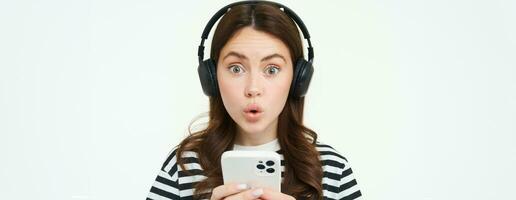 Portrait of happy young modern woman, female model in wireless headphones, smiling and looking at camera, holding smartphone, using mobile phone app, white background photo