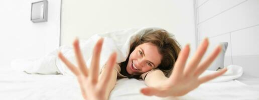Beautiful woman lying in bed, stretching her arms towards camera and laughing, waking up in morning in happy mood photo