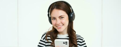 imagen de morena joven mujer, sonriente, escuchando a música en auriculares, acecho videos en móvil teléfono aplicación, participación teléfono inteligente, en pie terminado blanco antecedentes foto