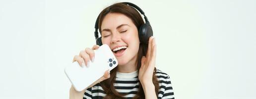 retrato de contento niña jugando karaoke aplicación, canto dentro teléfono inteligente micrófono, vistiendo inalámbrico auriculares, blanco antecedentes foto