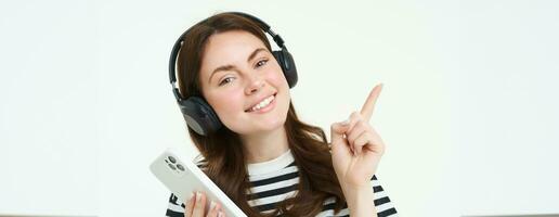 Portrait of smiling student, girl in headphones, holding mobile phone, pointing left, showing advertisement, store offer, white background photo