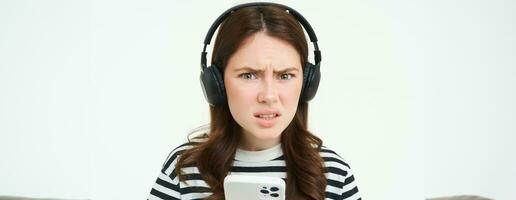 Image of puzzled young woman in headphones, holds smartphone, frowning and looking confused at camera, standing over white background photo