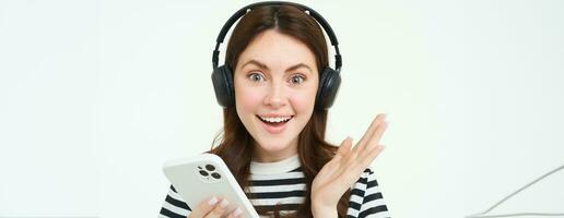 alegre, hermosa joven mujer utilizando teléfono inteligente aplicación, celebrando, mirando contento mientras jugando con móvil teléfono, escuchando música en inalámbrico auriculares foto