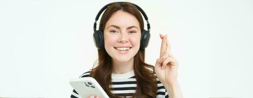 Portrait of smiling female model using headphones and smartphone, hold fingers crossed, makes wish, hopes for something, anticipates, stands over white background photo