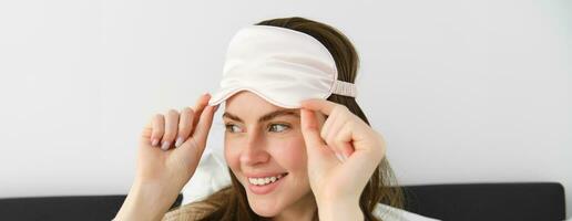 Portrait of smiling young modern woman open her eyes in morning, waking up and taking off sleeping mask, looking outside window, resting in bed, lying in bedroom photo