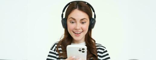 Young woman with surprised face, looking at smartphone screen, reacts amazed, looks with happiness at mobile phone, standing in wireless headphones, white background photo