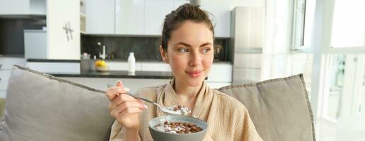 relajante Mañana a hogar. joven hermosa mujer en bata de baño, comiendo cereales en sofá, teniendo su desayuno en vivo habitación foto