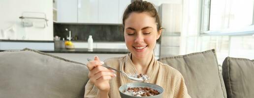 Portrait of gorgeous brunette woman sits on sofa, having her breakfast at home, smiling while eating cereals, holding bowl and spoon and laughing photo