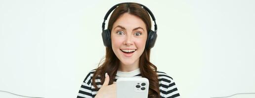 Young woman with surprised face, looking at smartphone screen, reacts amazed, looks with happiness at mobile phone, standing in wireless headphones, white background photo