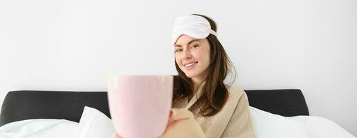 hermosa sonriente moderno mujer en pijama y dormido mascarilla, ofrecimiento taza de té o café en cama y mirando feliz, sentado en su dormitorio y relajante foto